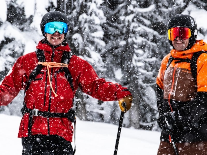 Revelstoke Mountain Resort opening day 2025 guests, two of them in ski gear and holding ski poles as they look at the camera and smile. Snow is noticeably falling.