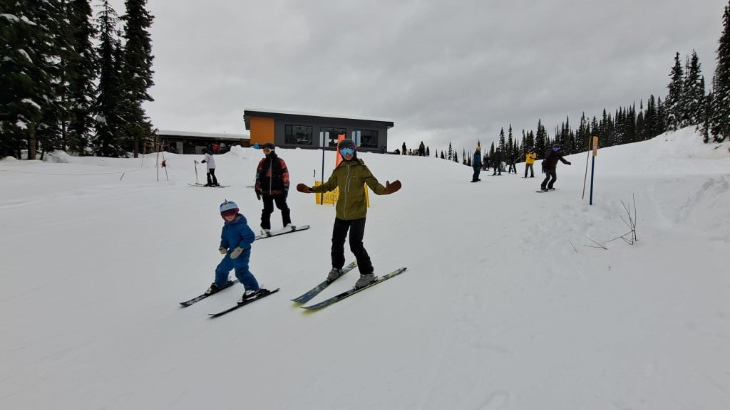 Two skiers, one adult and one child, and a snowboarder head down a trail while waving at the camera. 