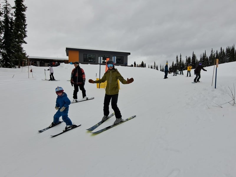 Two skiers, one adult and one child, and a snowboarder head down a trail while waving at the camera.