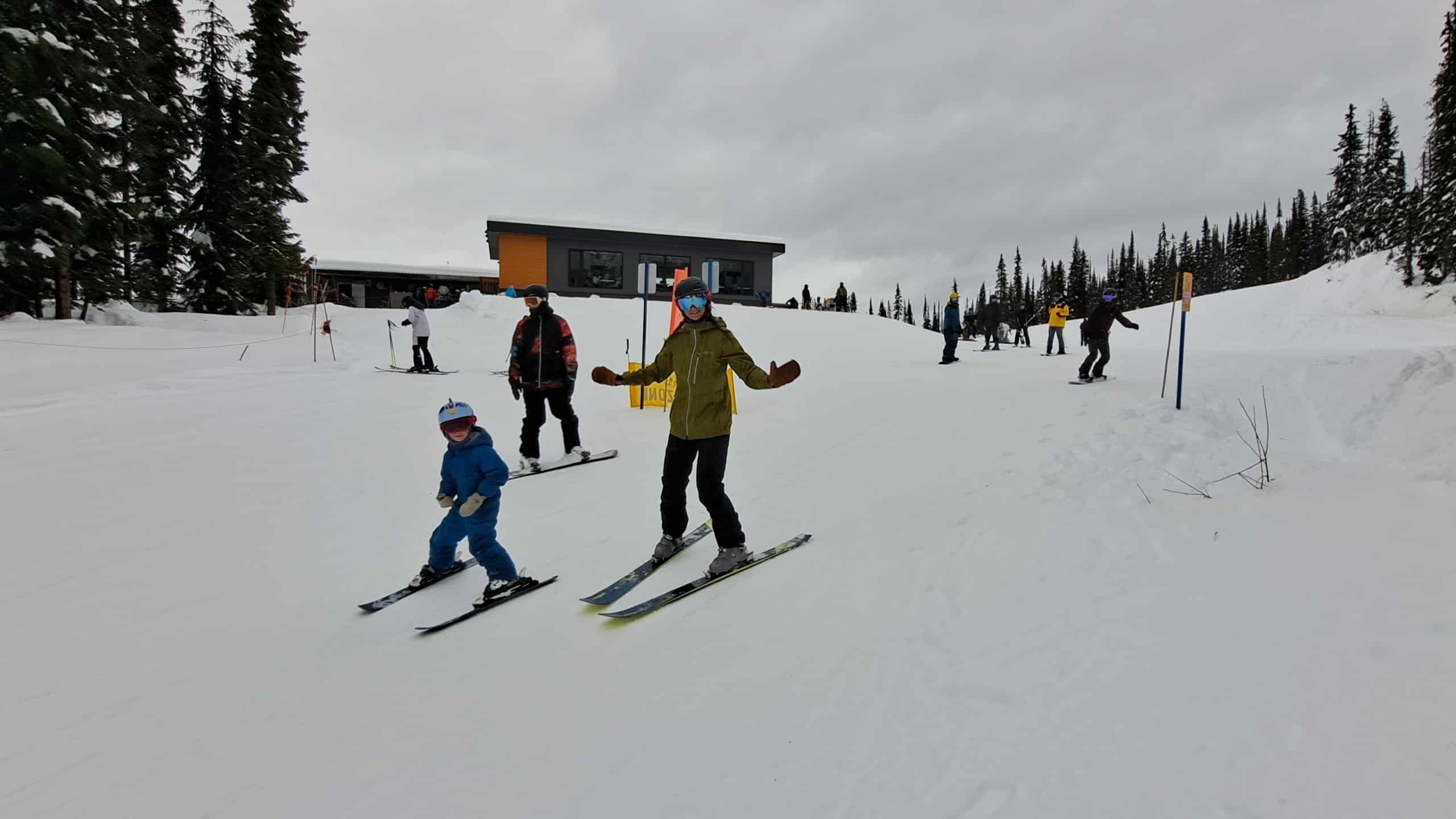 Two skiers, one adult and one child, and a snowboarder head down a trail while waving at the camera.
