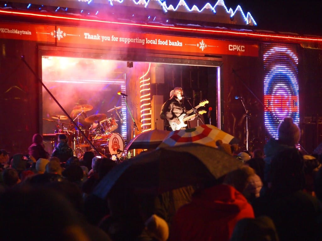 Ed Robertson, Barenaked Ladies lead singer and guitarist on the Hiliday Train 2026 stage looking out over a crowd in the dark. The stage and train cars are decorated with holiday lights.