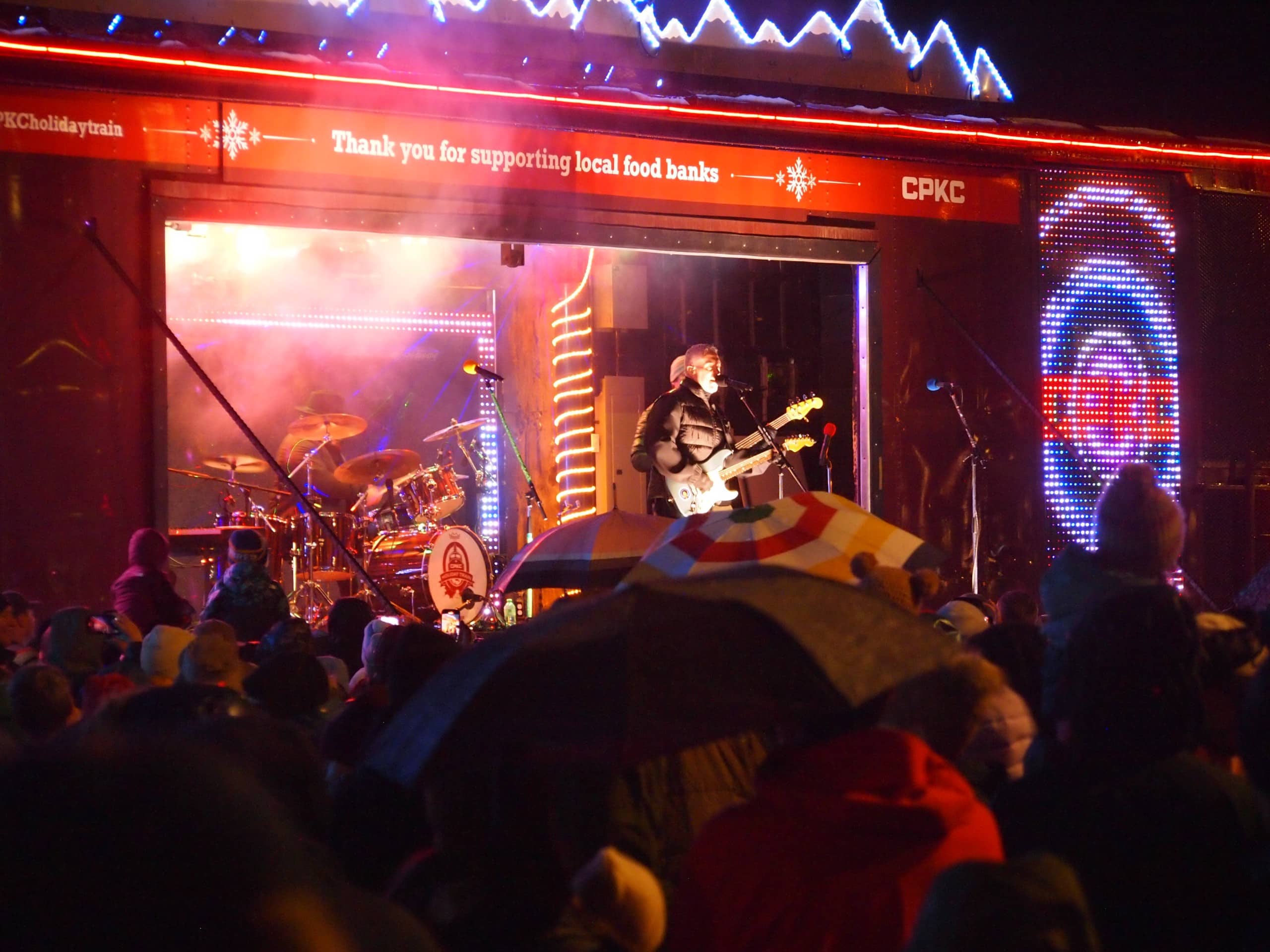 Ed Robertson, Barenaked Ladies lead singer and guitarist on the Hiliday Train 2026 stage looking out over a crowd in the dark. The stage and train cars are decorated with holiday lights.