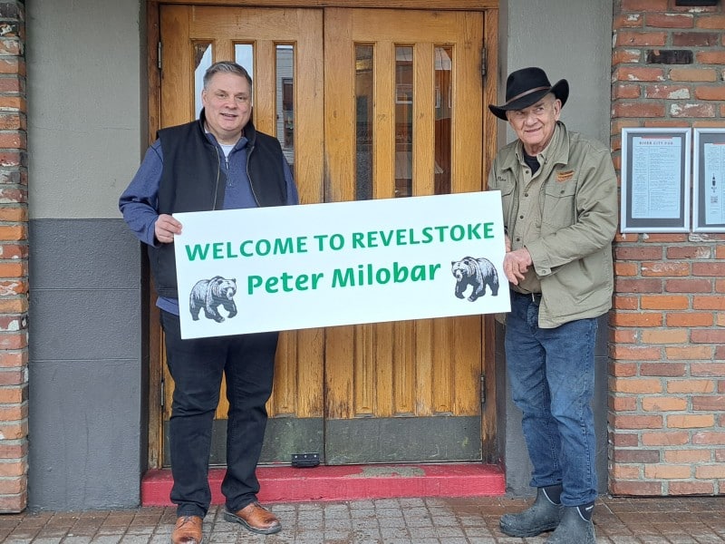Peter Milobar and Peter Bernaki in front of the Regent hotel holding a sign that reads, "Welcome to Revelstoke, Peter Milobar."