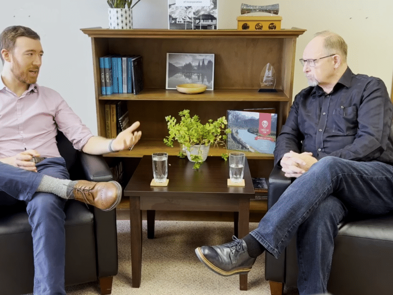 Paul Simon, a younger white man and Gary Sulz, and older white man sitting in chairs partially facing each other in an office and talking about Revelstoke STRs