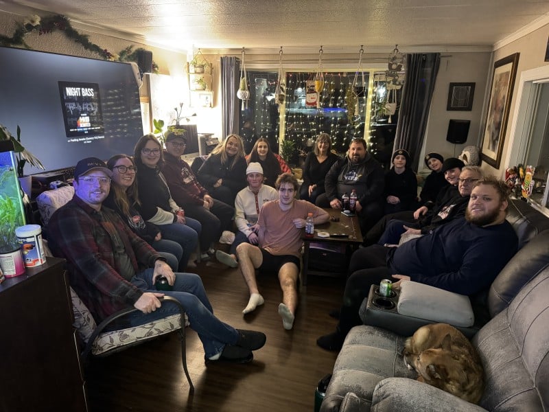 Revelstoke Grizzlies and their billet families in a large living room, a seated in different couches and chairs as they look at the camera and smile.