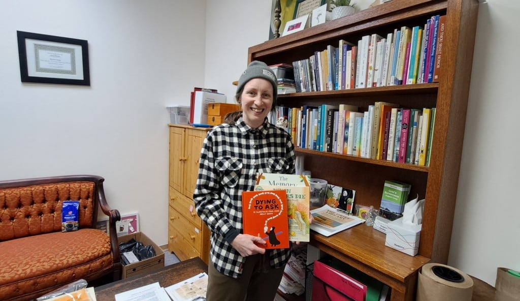 Theresa Hamilton, Revelstoke Hospice Society executive director standing in front of a book shelf filled with books. She is holding three children's books on the topic of death and grief.