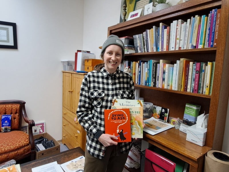 Theresa Hamilton, Revelstoke Hospice Society executive director standing in front of a book shelf filled with books. She is holding three children's books on the topic of death and grief.