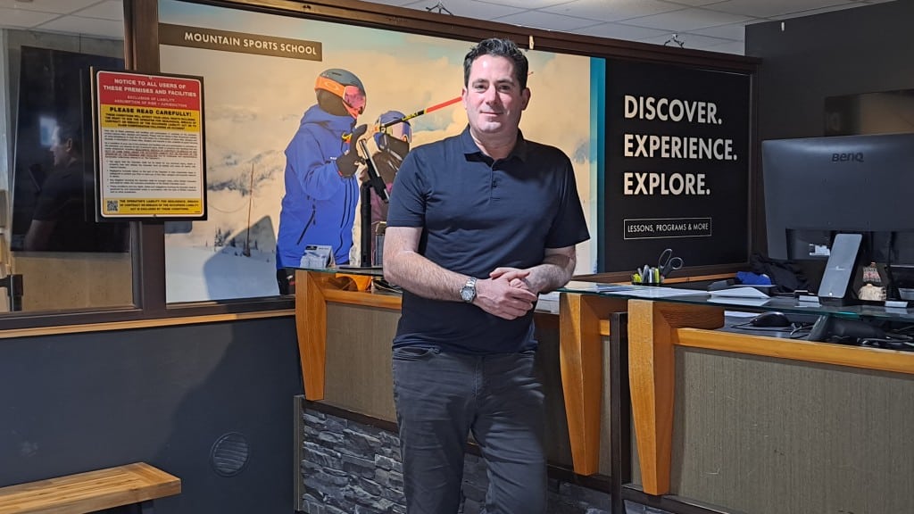 Jason Kelder, a middle-aged white man leaning against a desk at Revelstoke Mountain Resort's guest services. He is looking at the camera and smiling.