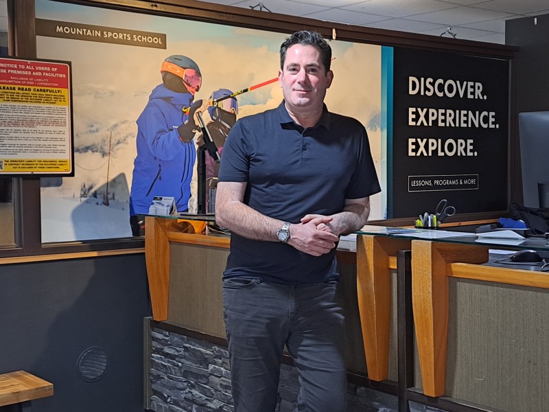 Jason Kelder, a middle-aged white man leaning against a desk at Revelstoke Mountain Resort's guest services. He is looking at the camera and smiling.