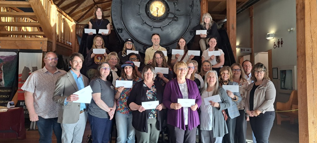 Over 20 Revelstoke Community Foundation Spring grant recipients stand in front of the Revelstoke Railway Museum train, all holding up white envelopes and looking at the camera smiling.