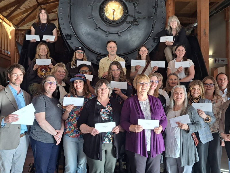 Over 20 Revelstoke Community Foundation Spring grant recipients stand in front of the Revelstoke Railway Museum train, all holding up white envelopes and looking at the camera smiling.