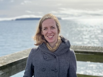 The Fall-Down Effect author Liz Johnston standing on a dock overlooking a large body of water. Her back is to the water, she is looking at the camera and smiling.