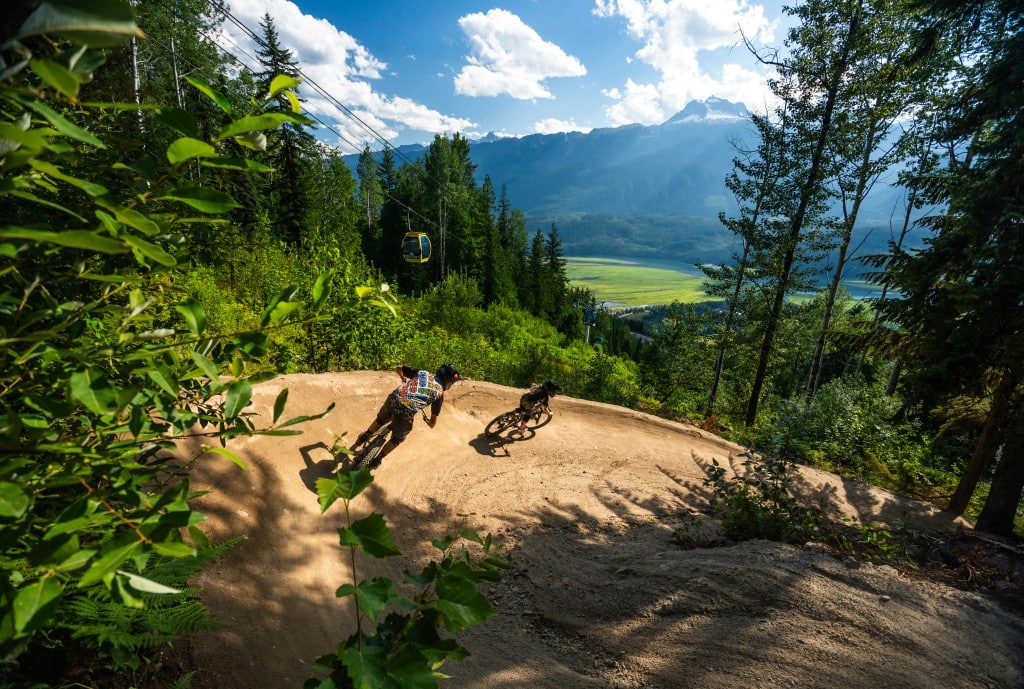 Two bikers on a dirt trail at Revelstoke Mountain Resort with a gap in the trees showing the lower valley and one of the resort gondola cars