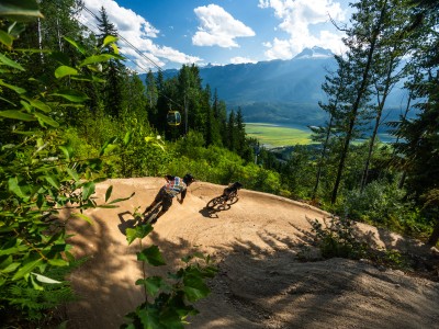 Two bikers on a dirt trail at Revelstoke Mountain Resort with a gap in the trees showing the lower valley and one of the resort gondola cars
