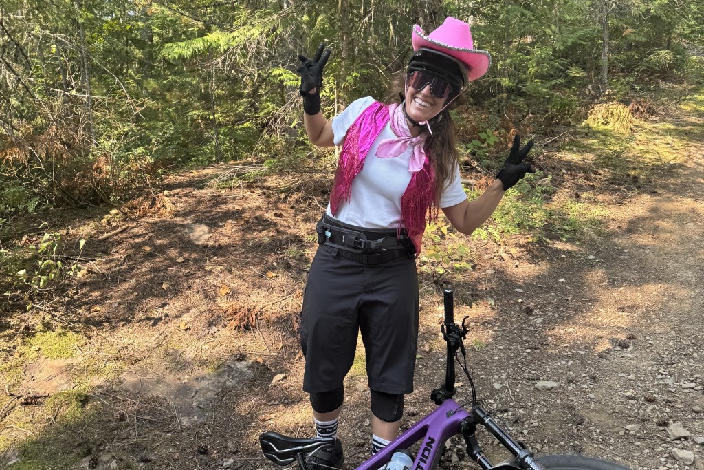 Meghan Tabor in a pink cowboy hat, biking gear with a mountain bike at her feet. She is looking at the camera while smiling and throwing peace signs.