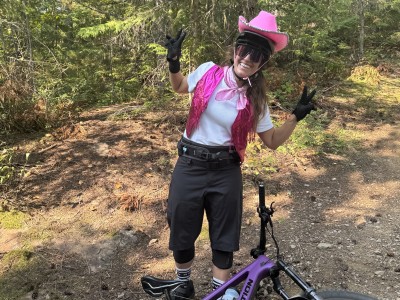 Meghan Tabor in a pink cowboy hat, biking gear with a mountain bike at her feet. She is looking at the camera while smiling and throwing peace signs.