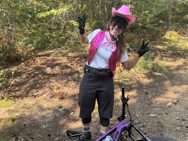 Meghan Tabor in a pink cowboy hat, biking gear with a mountain bike at her feet. She is looking at the camera while smiling and throwing peace signs.