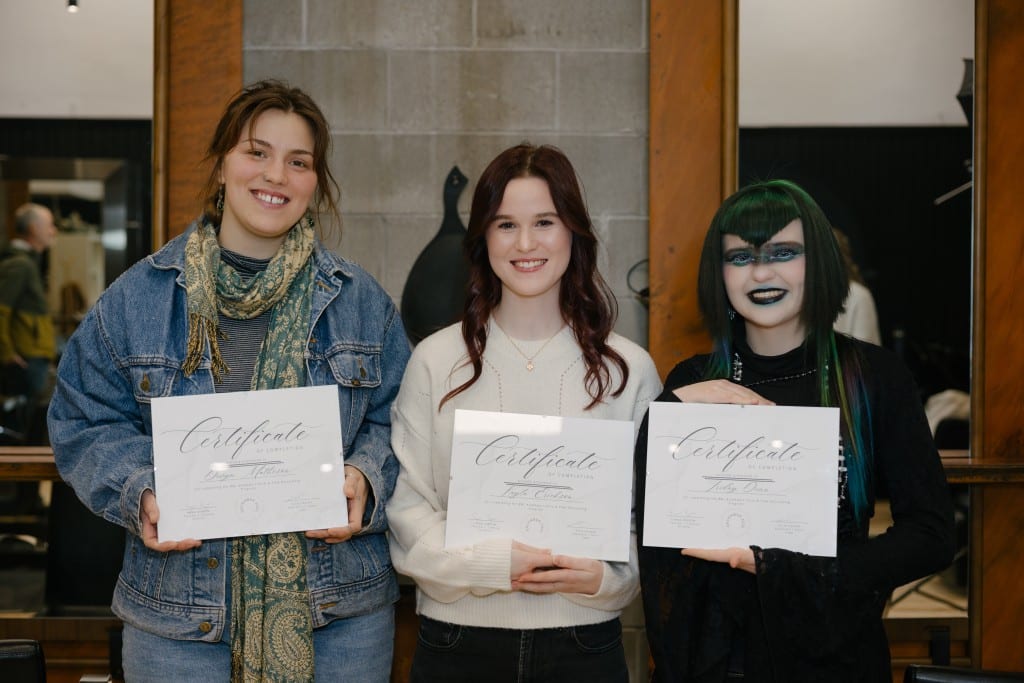 Three Birch and Lace hair stylist academy program graduates stand side by side holding their certificates in front of them. All three are looking at the camera and smiling.