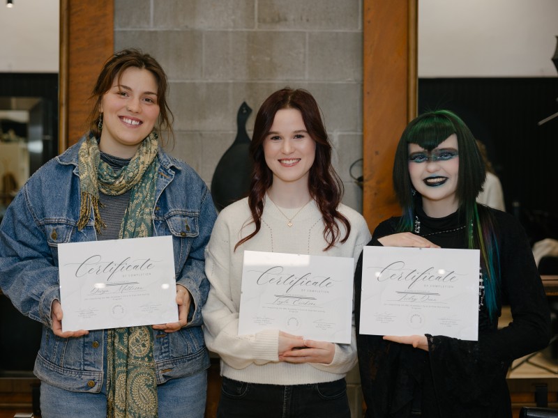 Three Birch and Lace hair stylist academy program graduates stand side by side holding their certificates in front of them. All three are looking at the camera and smiling.