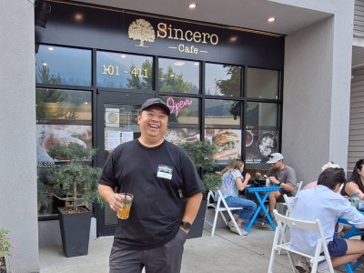 Andy Nguyen, a philipino man stands in front of his cafe, Sincero. He is holding an orange drink and laughing as he looks past the camera. Behind him are customers at the outdoor seating. Photo is after his interview about Revelstoke commercial spaces.