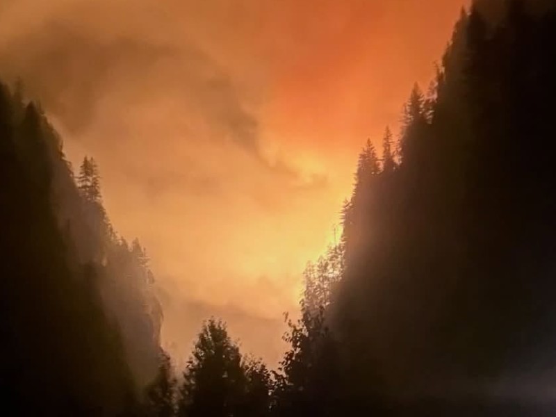 A night photo of the Incomappleux River Fish River wildfire that prompted an evacuation order from City of Revelstoke. The forest, lake and valley are a nighttime black, and the center of the photo where the valley meets is orange and smokey from fire.