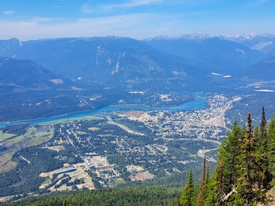 A wide view of the City of Revelstoke, Columbia River and the valley that the city resides in. Trees are green and the slightly cloudy sky is also a bit hazy, indicative of the wildfires in the region at the time of the photo.