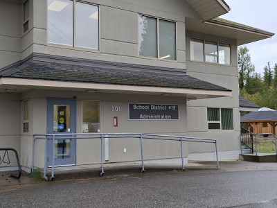 External shot of Revelstoke school district 19 administration building, a two-story, cream coloured building with grey tiling on the roof and front entrance overhang. There is a concrete ramp leading up to the front door and a sign that reads School District #19 administration